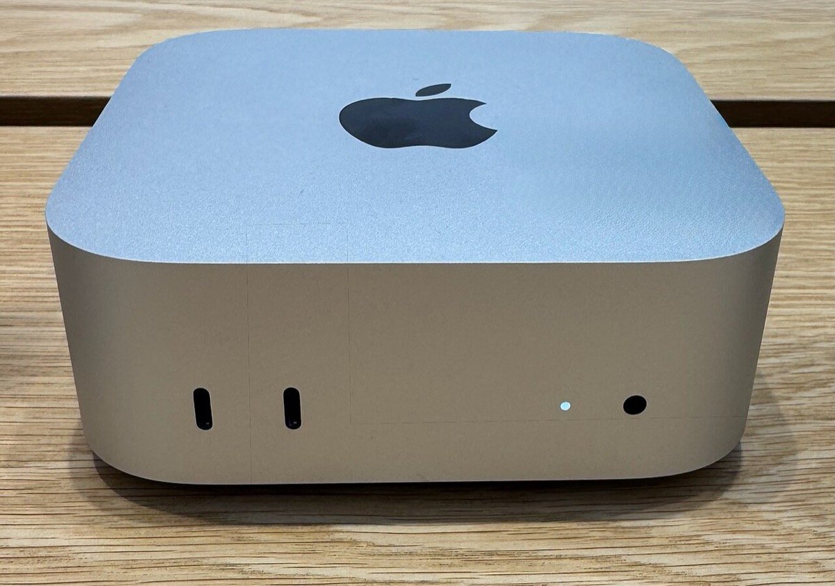 A silver Mac mini on a wooden desk, seen from the front, with two USB-C ports and a headphone socket